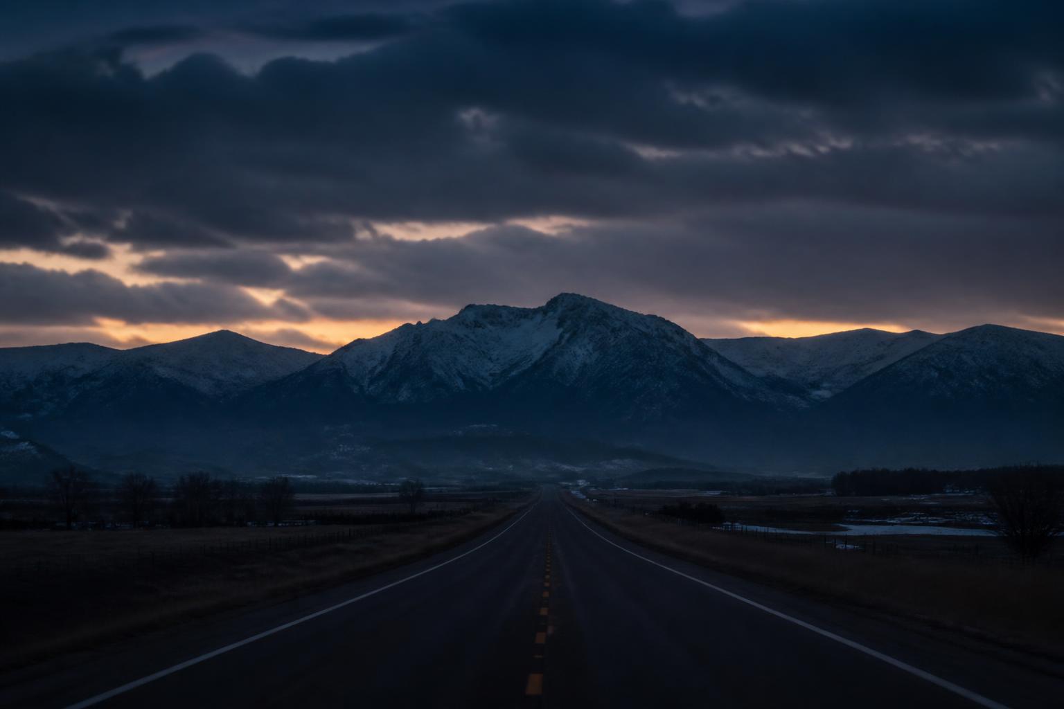 Painted illustration of an empty two-lane county road running west through Northern Colorado farmland at last light with the Front Range on the horizon.