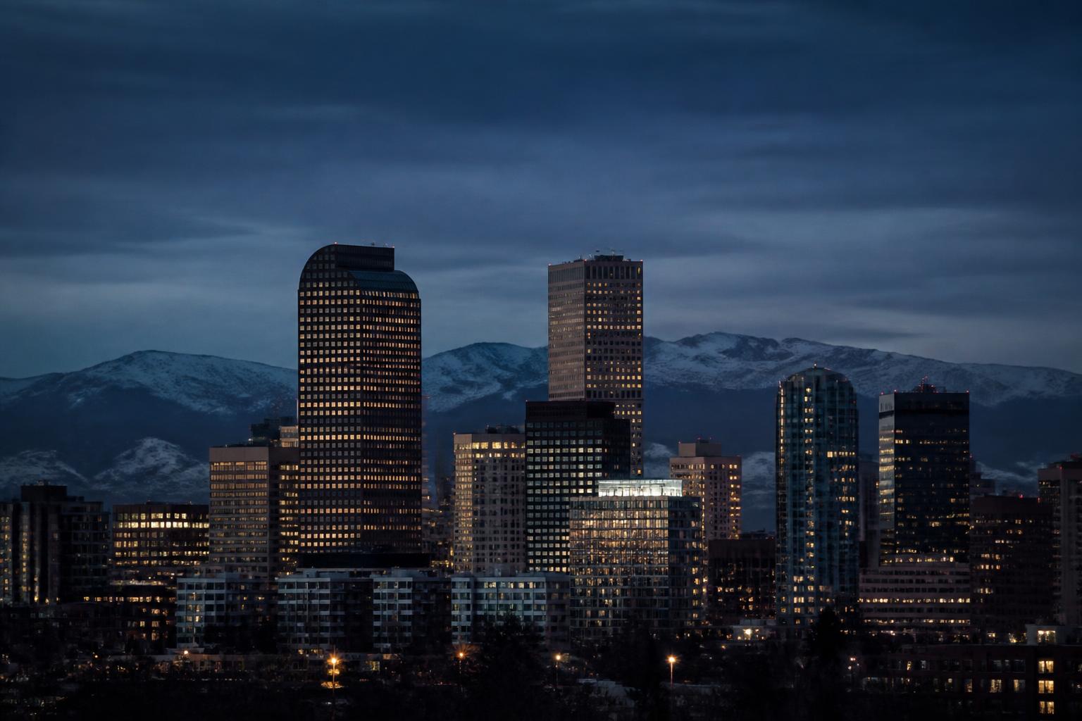 Painted illustration of the Denver, Colorado skyline at dusk with a lit clock tower among the towers and the Rocky Mountains behind in deep violet.